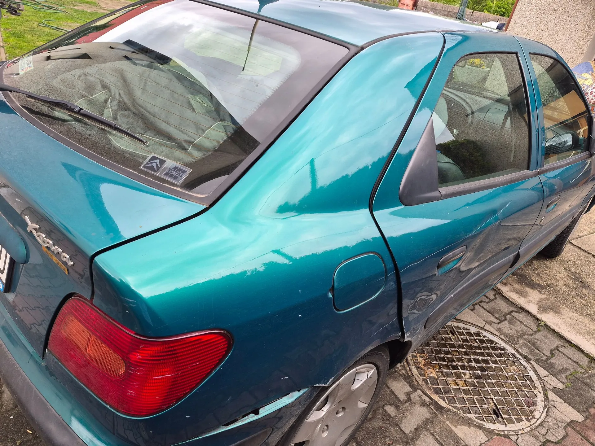 Tylna część samochodu marki Citroën w kolorze turkusowym. Widoczny jest lewy tylny błotnik z wgnieceniami i zarysowaniami, tylna lampa oraz fragment klapy z napisem 'Xantia'. Samochód zaparkowany jest na kostce brukowej obok metalowego włazu kanalizacyjnego.