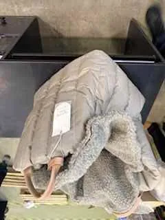 Beżowa pikowana kurtka z futrzanym kołnierzem leżąca na czarnym stole. Kurtka posiada białą etykietę. W tle widoczne są drewniane deski i betonowa podłoga. W prawym dolnym rogu obrazu znajduje się fragment czarnej buty.