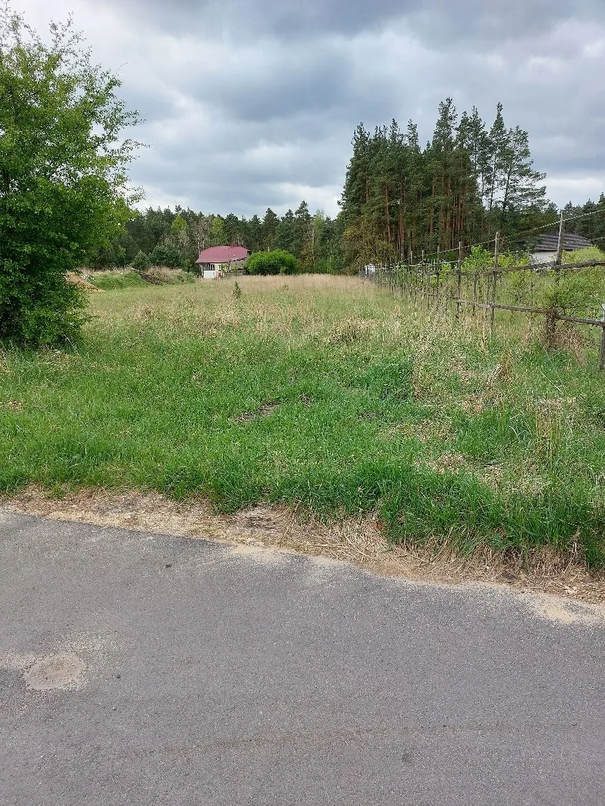 Krajobraz wiejski z widokiem na trawiaste pole. Na pierwszym planie znajduje się fragment asfaltowej drogi. Po prawej stronie biegnie drewniany płot, a w oddali widoczny jest biały dom z czerwonym dachem oraz gęsty las sosnowy pod zachmurzonym niebem.