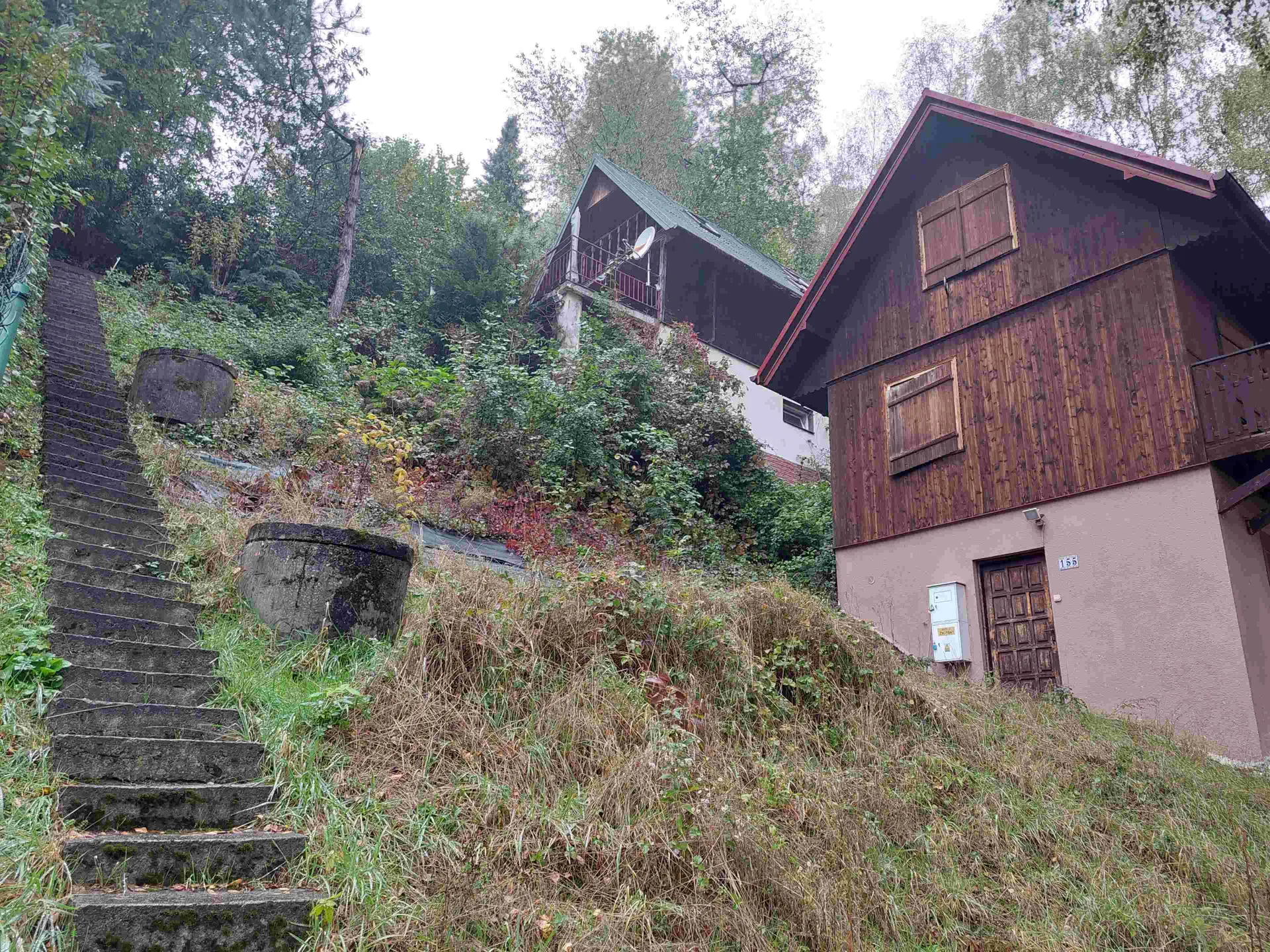 Widok na stromy zbocze z kamiennymi schodami po lewej stronie, porośniętymi mchem. Na zboczu znajdują się dwa duże betonowe, cylindryczne zbiorniki. W tle widoczne są dwa domy: lewy z zielonym dachem, balkonem i anteną satelitarną, prawy z czerwonym dachem, brązową drewnianą elewacją na górze i różową ścianą na dole. Przy drzwiach prawego domu umieszczono tabliczkę z numerem 155 oraz białą skrzynkę. Całość otoczona jest gęstym lasem i wysoką trawą. Niebo jest pochmurne.