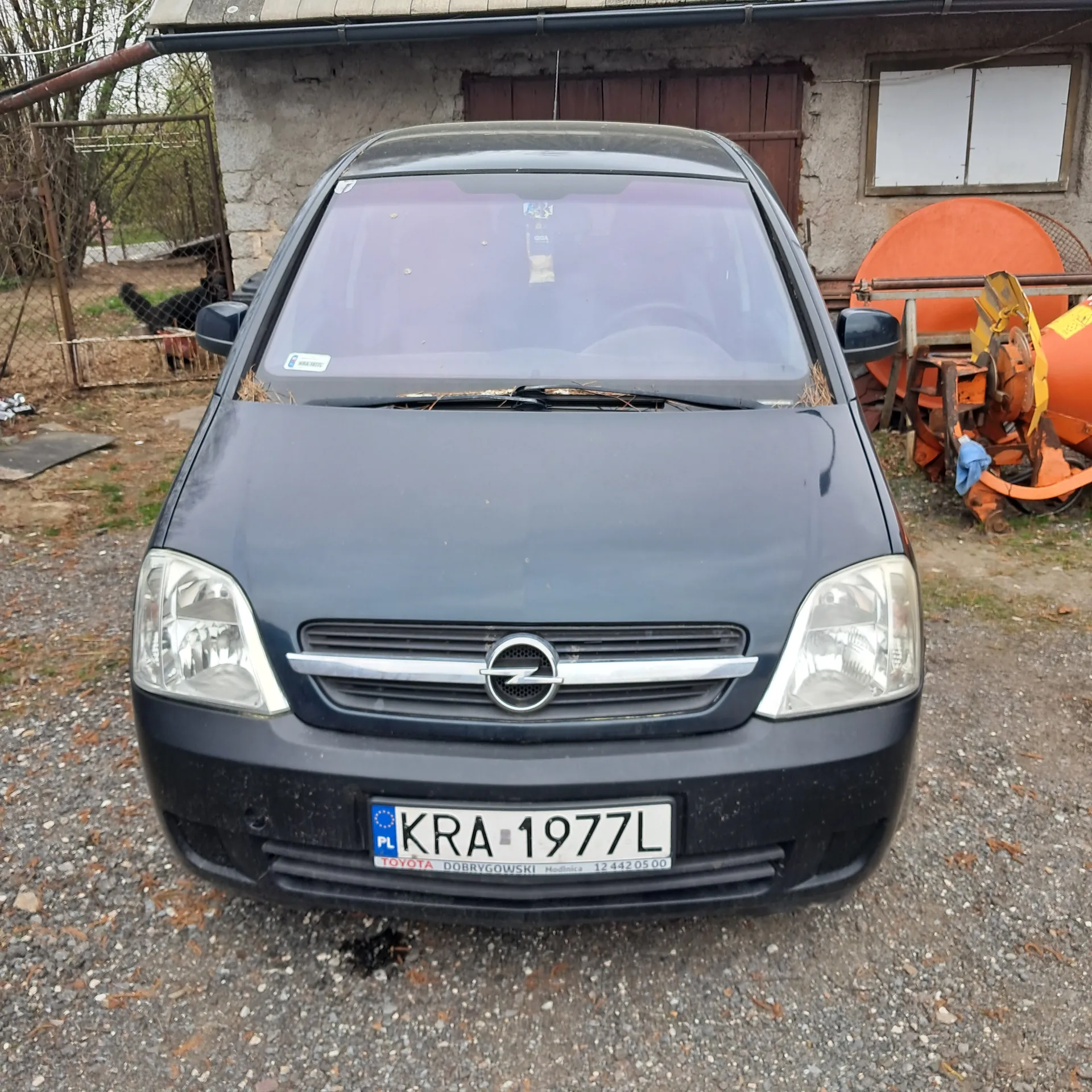 Ciemnoszary samochód marki Opel z przodu, z widoczną tablicą rejestracyjną KRA 1977L. Na tablicy widoczne są również napisy 'PL', 'TOYOTA DOBRYGOWSKI' oraz numer '12 442 05 00'. W tle budynek z oknem, pomarańczowe maszyny rolnicze i żwirowa powierzchnia.