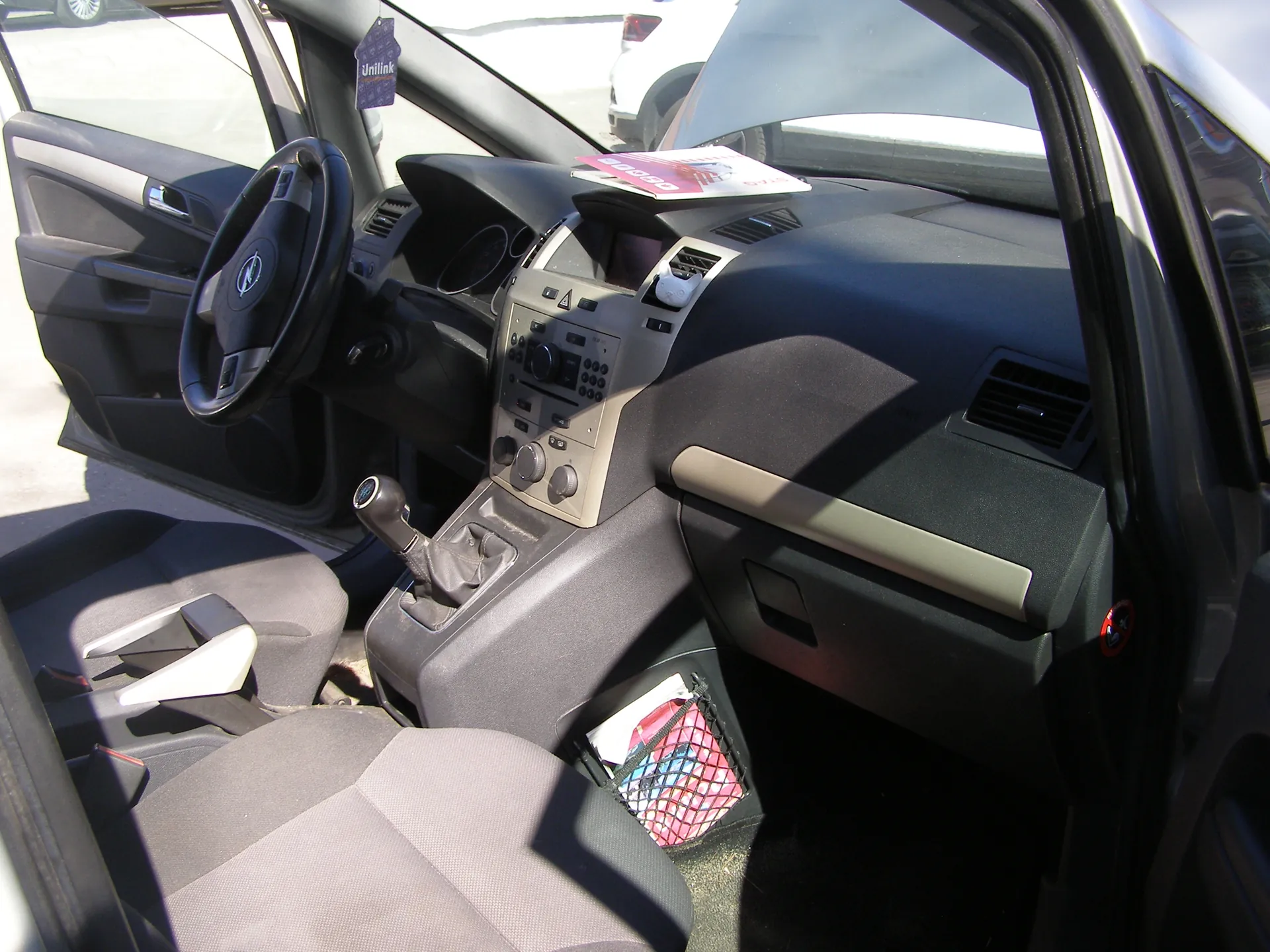 Wnętrze samochodu z otwartą lewą przodem drzwiami. Widoczna kierownica z logo, deska rozdzielcza z systemem audio i klimatyzacją, dźwignia zmiany biegów oraz szare siedzenia. Na desce rozdzielczej leży różowo-biały dokument. Na lusterku wstecznym wiszący niebieski znaczek z napisem "Unlink". W siatce na prawej stronie pasażyra znajdują się przedmioty, w tym kolorowy element w kratkę. Poza samochodem widoczne są inne pojazdy i asfalt.