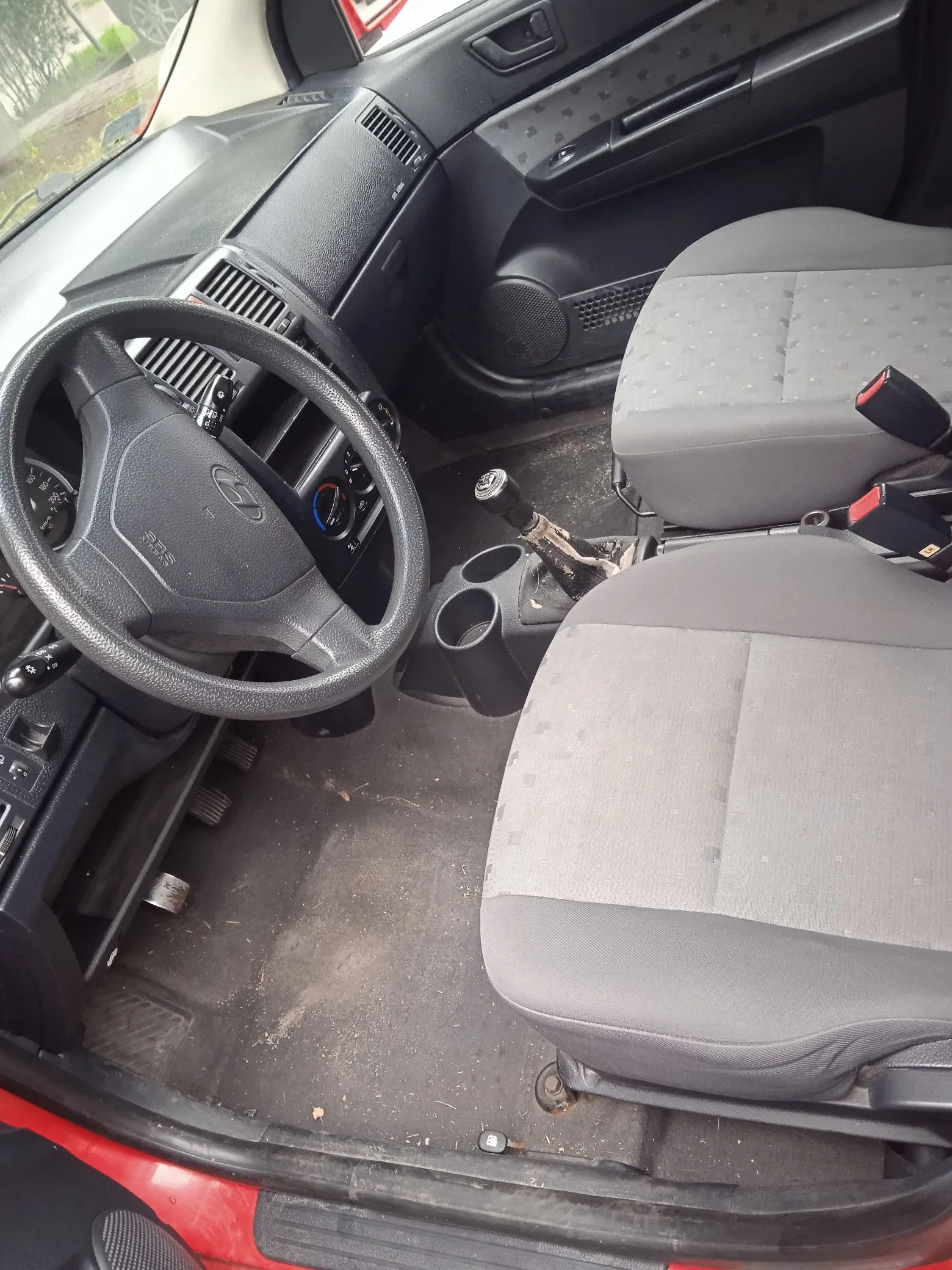 Wnętrze samochodu widziane z perspektywy otwartych drzwi pasażera. Widoczna jest kierownica z logo Hyundai, deska rozdzielcza z nawiewami, dwa szare materiałowe fotele, lewarek zmiany biegów oraz podłoga z dywanikami. Samochód ma czerwony kolor nadwozia widoczny na progu.
