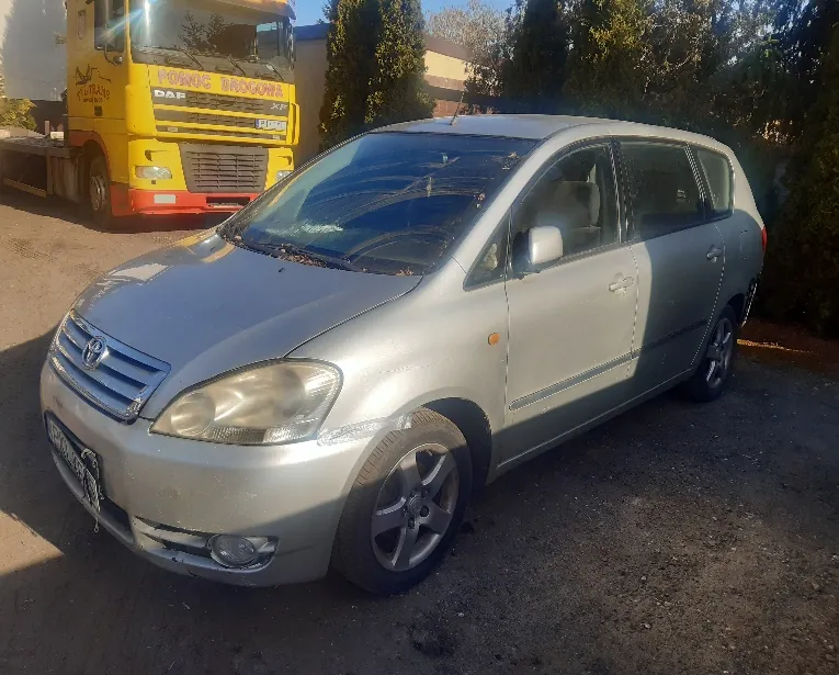 Srebrny samochód osobowy typu minivan zaparkowany na podwórku. W tle żółty ciężarówka z napisem "POMOC DROGOWA" oraz logo "DAF" na masce. Widoczne drzewa, budynki i nierówna powierzchnia podłogi. Tablica rejestracyjna samochodu jest częściowo widoczna.