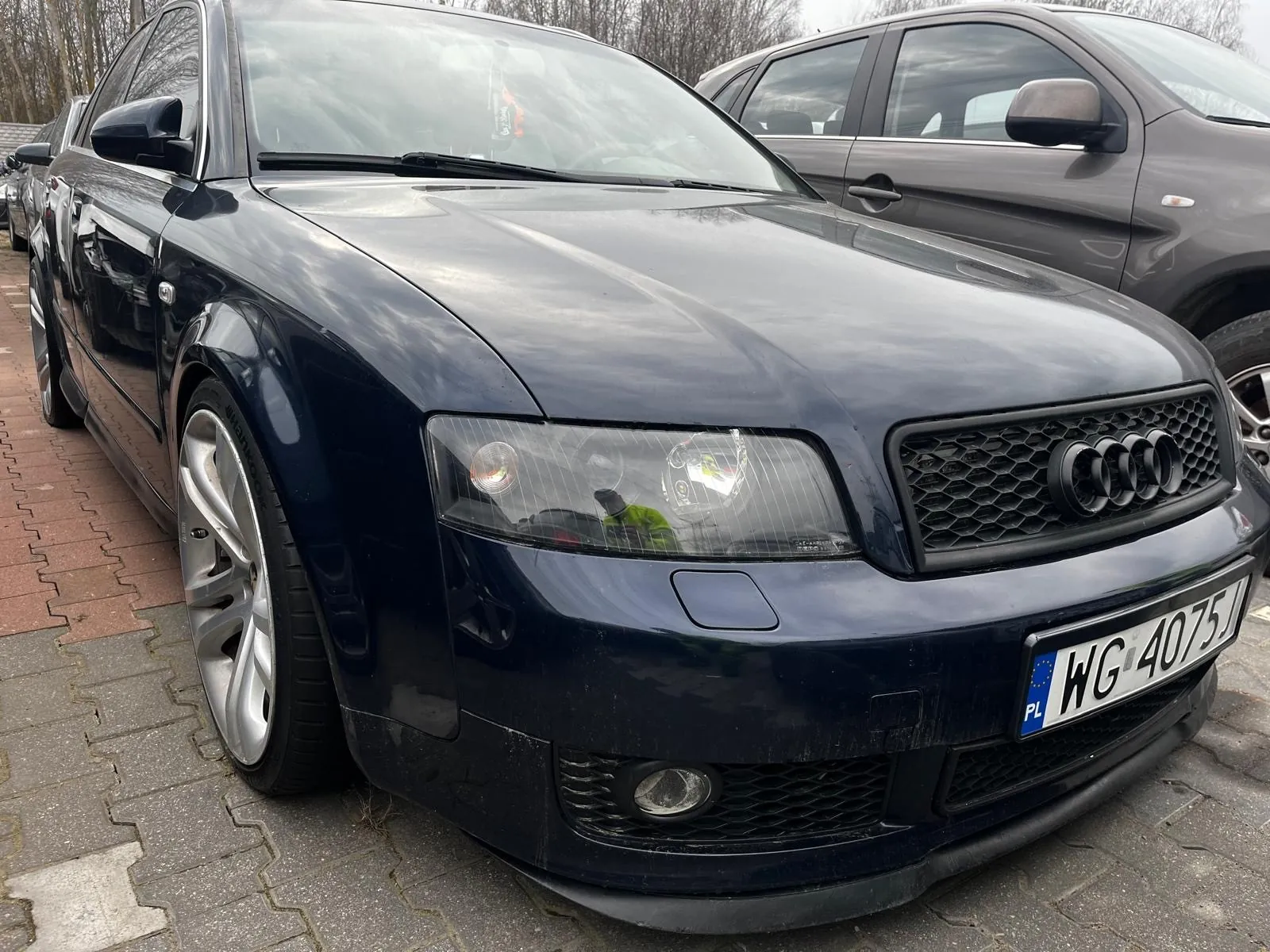 Czarny samochód marki Audi zaparkowany na brukowanej powierzchni. Widoczna przód pojazdu z charakterystyczną atrapą chłodnicy z czterema pierścieniami, reflektorem i tablicą rejestracyjną o numerze WG 4075J z niebieskim pasem po lewej stronie zawierającym oznaczenie PL. W tle częściowo widoczny szary samochód oraz drzewa bez liści. Podłoga składa się z czerwono-szarych elementów brukowych.