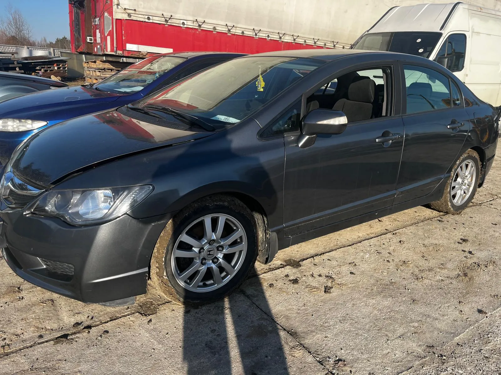 Ciemnoszary samochód osobowy typu sedan z widocznym uszkodzeniem na masce i brudnymi kołami, zaparkowany na betonowej powierzchni. W tle niebieski samochód, czerwono-biały naczepa ciężarowa oraz biały furgon. Na przedniej szybie ciemnoszarego samochodu widoczny żółty znaczek. Tło obejmuje również stosy materiałów i drzewa pod jasnym niebem.