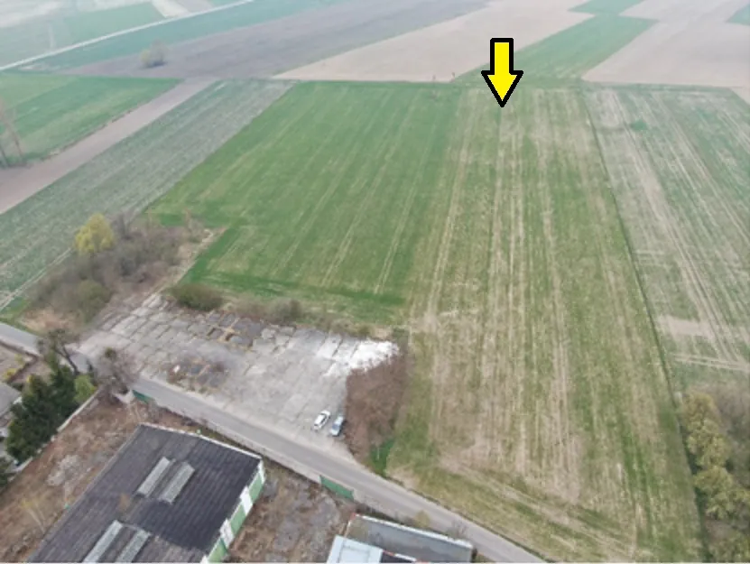 Zdjęcie lotnicze przedstawiające obszar rolniczy z polami w różnych odcieniach zieleni i brązu. W dolnej części obrazu widoczny jest betonowy plac z zaparkowanymi dwoma samochodami oraz fragmenty budynków gospodarczych. W górnej części zdjęcia znajduje się żółta strzałka skierowana w dół, wskazująca na konkretny punkt na zielonym polu.