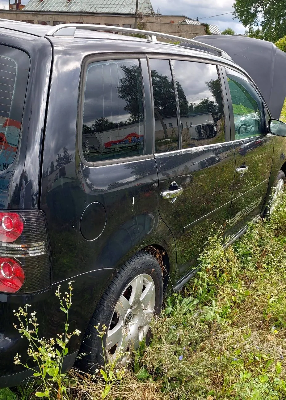 Boczny widok czarnego samochodu typu kombi zaparkowanego w wysokiej trawie. Widoczna jest tylna część nadwozia, tylne i boczne szyby, koło oraz uchwyty drzwi. W tle widać fragmenty budynków i drzewa.