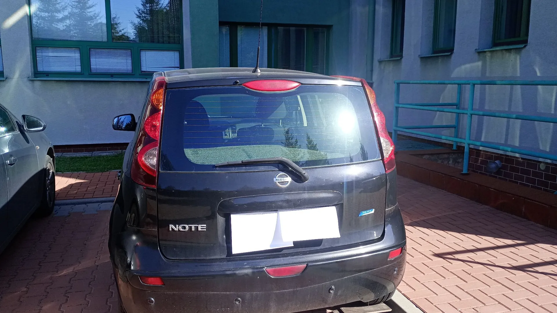 Tylny widok czarnego samochodu marki Nissan Note zaparkowanego na kostce brukowej przed budynkiem. Na klapie bagażnika widoczny jest napis NOTE oraz logo Nissana. Tablica rejestracyjna jest zakryta białym polem. W tle znajduje się jasny budynek z oknami i niebieską metalową barierką.