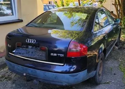 Tylny widok ciemnogranatowego samochodu marki Audi. Na klapie bagażnika widoczne jest logo Audi (cztery splecione pierścienie) oraz oznaczenie modelu '2.5 TDI'. Samochód nie posiada tablic rejestracyjnych, w ich miejscu znajduje się czarna ramka. Pojazd jest zaparkowany na nieutwardzonym podłożu obok jasnej ściany budynku.
