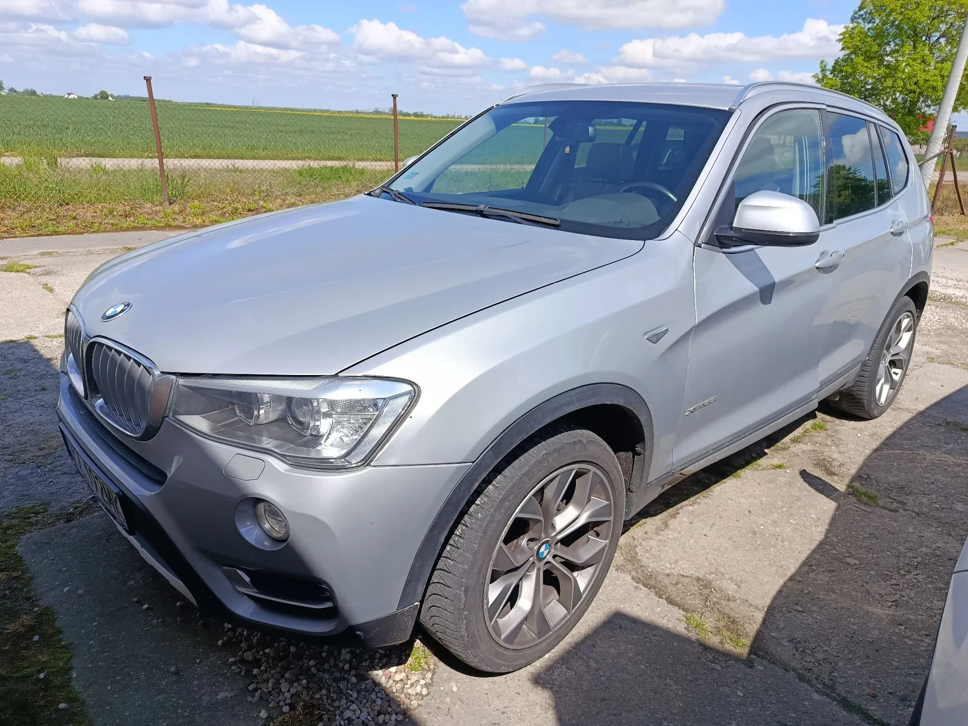 Srebrny samochód marki BMW typu SUV zaparkowany na betonowym podjeździe. Pojazd jest ukazany z przodu i z boku. W tle widać zielone pole oraz niebo z białymi chmurami.
