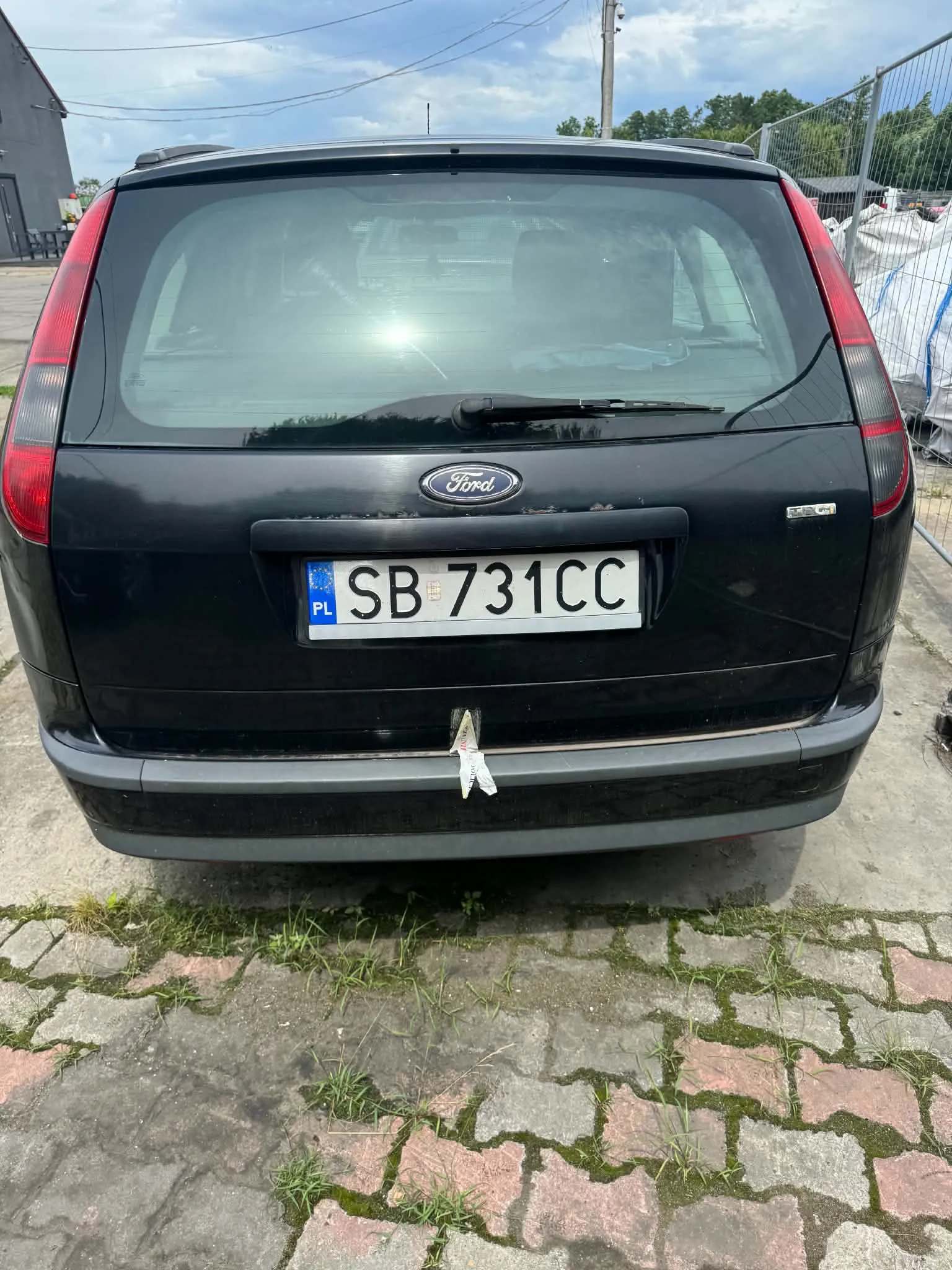 Tylny widok czarnego samochodu marki Ford. Na klapie bagażnika znajduje się logo Ford oraz polska tablica rejestracyjna o numerze SB 731CC. Samochód stoi na kostce brukowej z wyrastającą spomiędzy płyt trawą.