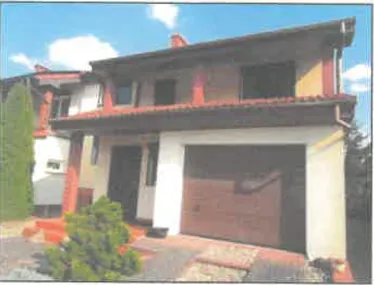 Zdjęcie dwukondygnacyjnego domu jednorodzinnego z garażem. Budynek ma jasną elewację z czerwonymi detalami architektonicznymi i ciemnym dachem. Na parterze znajduje się brązowa brama garażowa oraz wejście do domu. Przed domem widoczny jest podjazd oraz zielone krzewy i wysoki, wąski żywopłot po lewej stronie. W tle widać niebieskie niebo z białymi chmurami.