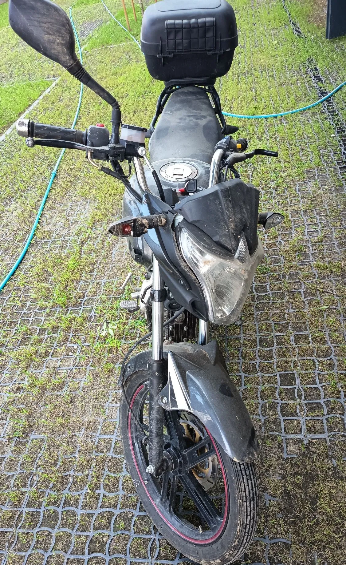 Czarny motocykl zaparkowany na plastikowej kratce trawnikowej. Motocykl jest brudny, posiada czarny kufer z tyłu oraz lusterko po lewej stronie. Widoczny jest reflektor przedni, kierownica z manetkami oraz przednie koło z czerwoną obwódką na feldze.