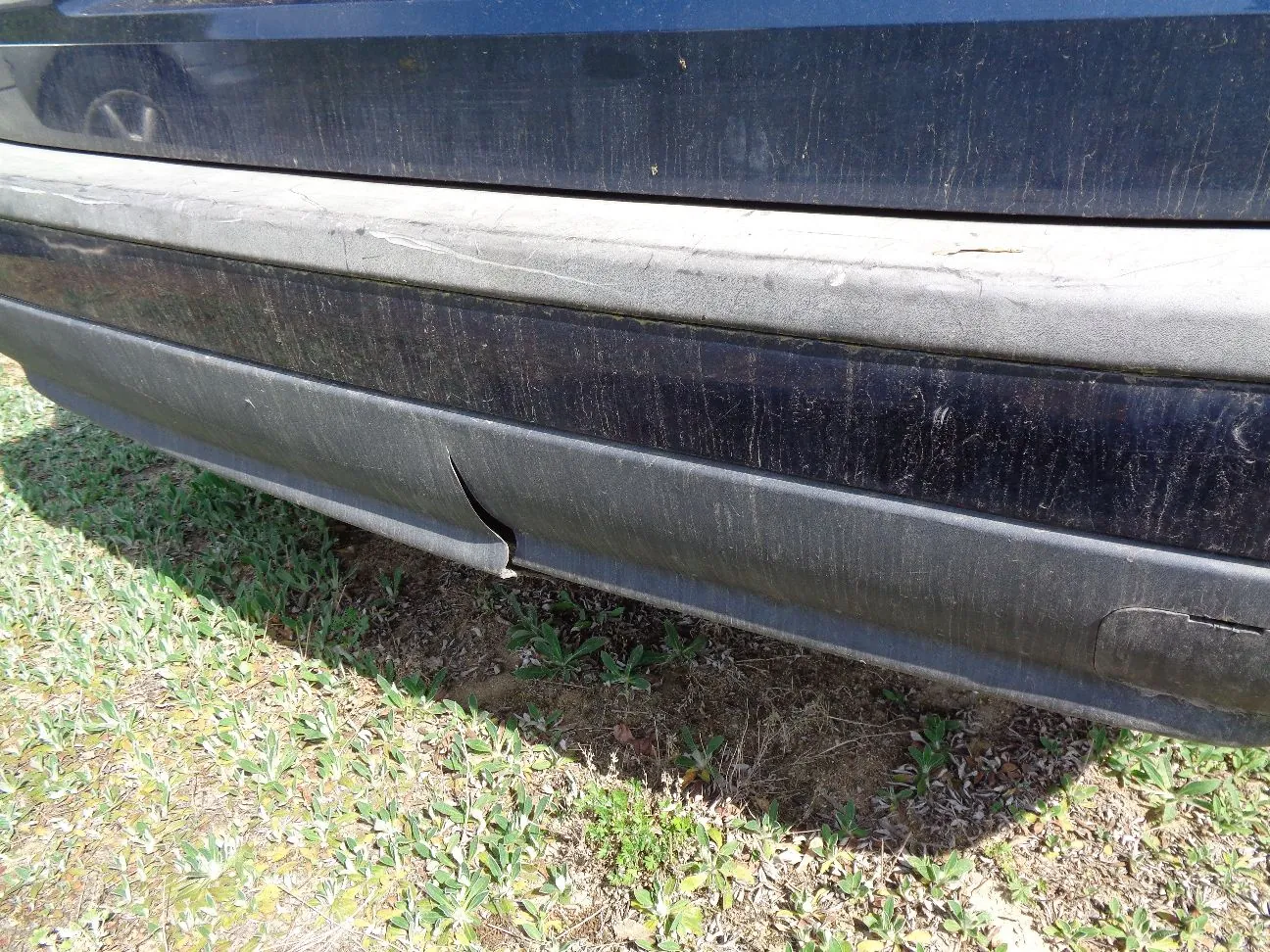 Zbliżenie na dolną część czarnego zderzaka samochodu. Plastikowy element jest porysowany i posiada widoczne wgniecenie oraz pęknięcie w dolnej części. Samochód stoi na trawiastym podłożu.