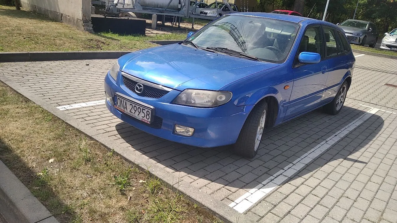 Niebieski samochód osobowy marki Mazda zaparkowany na kostce brukowej. Pojazd posiada polską tablicę rejestracyjną o numerze ONA 29958. W tle widoczny jest trawnik, krawężnik oraz inne zaparkowane samochody.