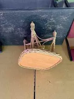 Mały stołek z plecionym siedziskiem i drewnianymi nogami umieszczony na ciemnej, zużytej powierzchni. W tle widoczna beżowa ściana i fragment różowego przedmiotu.