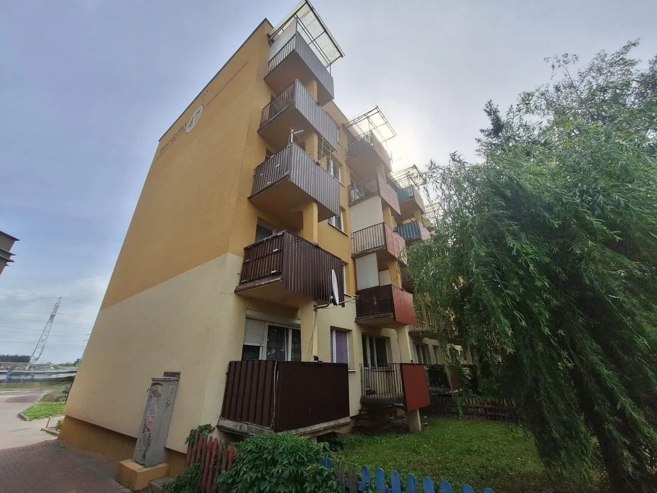 Wielopiętrowy budynek mieszkalny o żółtej elewacji z licznymi balkonami z brązowymi balustradami. Po prawej stronie zdjęcia znajduje się duże, gęste zielone drzewo, które częściowo zasłania budynek. Przed budynkiem widoczny jest trawnik oraz fragment niebieskiego płotu.