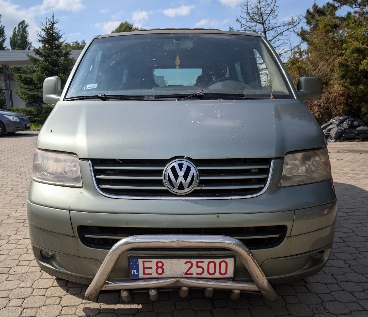 Widok z przodu srebrno-szarego vana marki Volkswagen. Pojazd posiada zamontowaną srebrną rurę ochronną (bullbar) na przednim zderzaku, do której przymocowana jest tablica rejestracyjna o numerze E8 2500 z oznaczeniem PL. Samochód stoi na kostce brukowej w słoneczny dzień.
