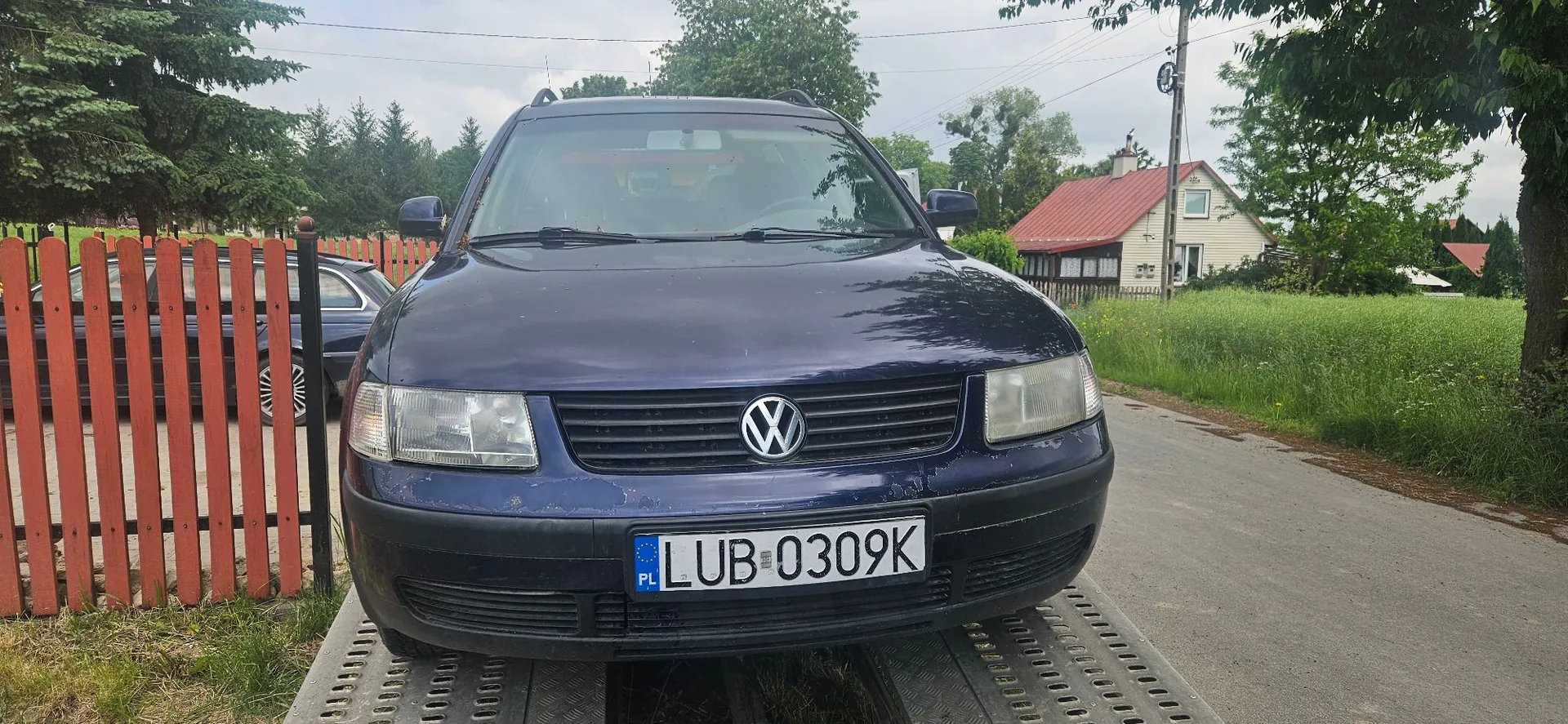 Granatowy samochód marki Volkswagen zaparkowany na metalowej platformie transportowej. Pojazd posiada polską tablicę rejestracyjną o numerze LUB 0309K. W tle widoczny jest czerwony płot, zielone drzewa oraz dom z czerwonym dachem.