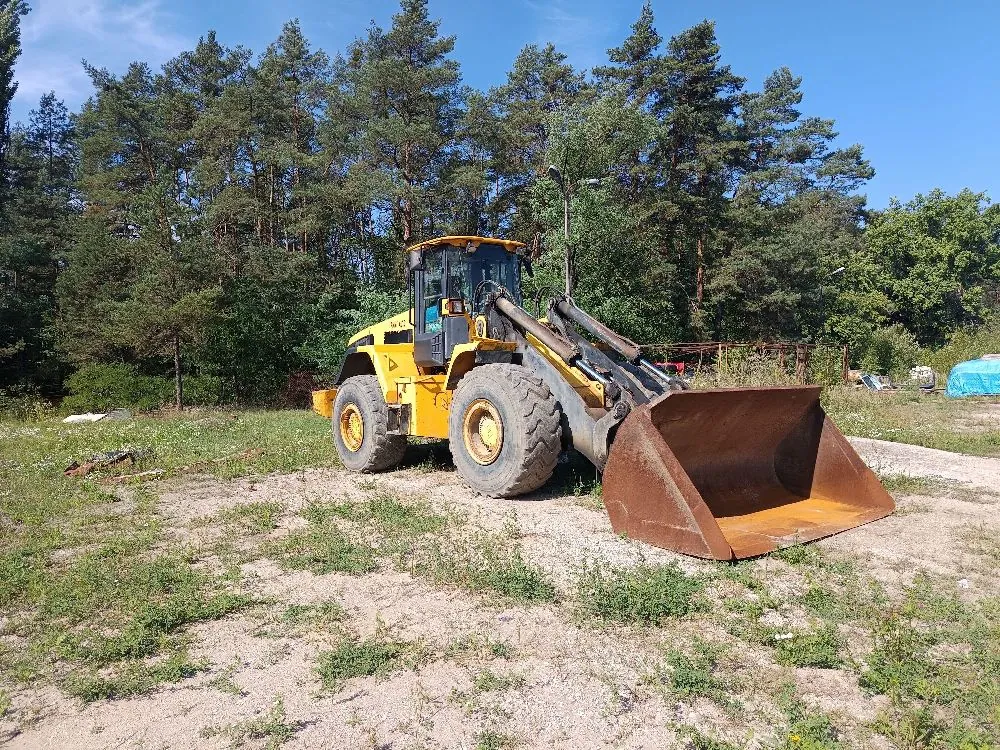Żółty ładowarka kołowa z dużym, brązowym łyżką z przodu, zaparkowana na piaszczystym terenie z niską roślinnością. W tle znajduje się gęsty las sosnowy pod bezchmurnym, niebieskim niebem.