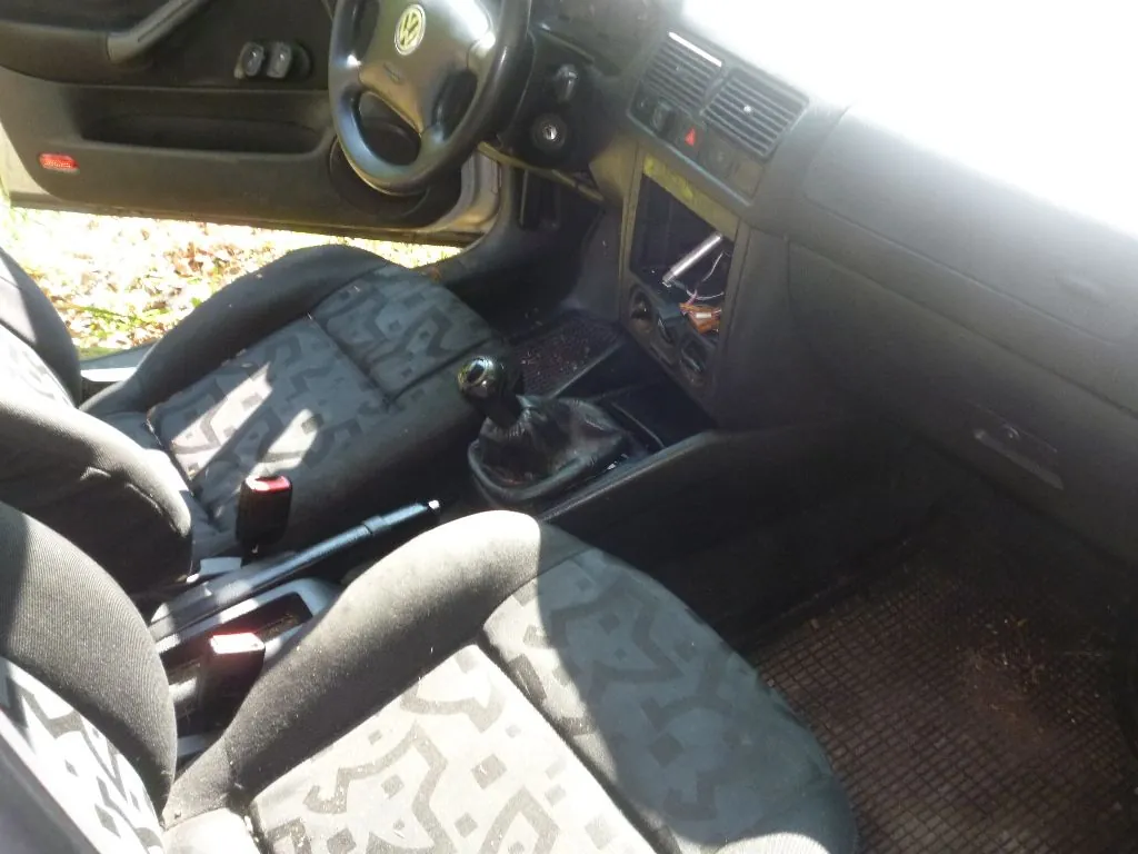 Wnętrze samochodu marki Volkswagen z perspektywy pasażera. Widoczna jest kierownica z logo VW, deska rozdzielcza z radiem, lewarek zmiany biegów oraz dwa przednie fotele z ciemną, wzorzystą tapicerką. Na podłodze znajdują się gumowe dywaniki.