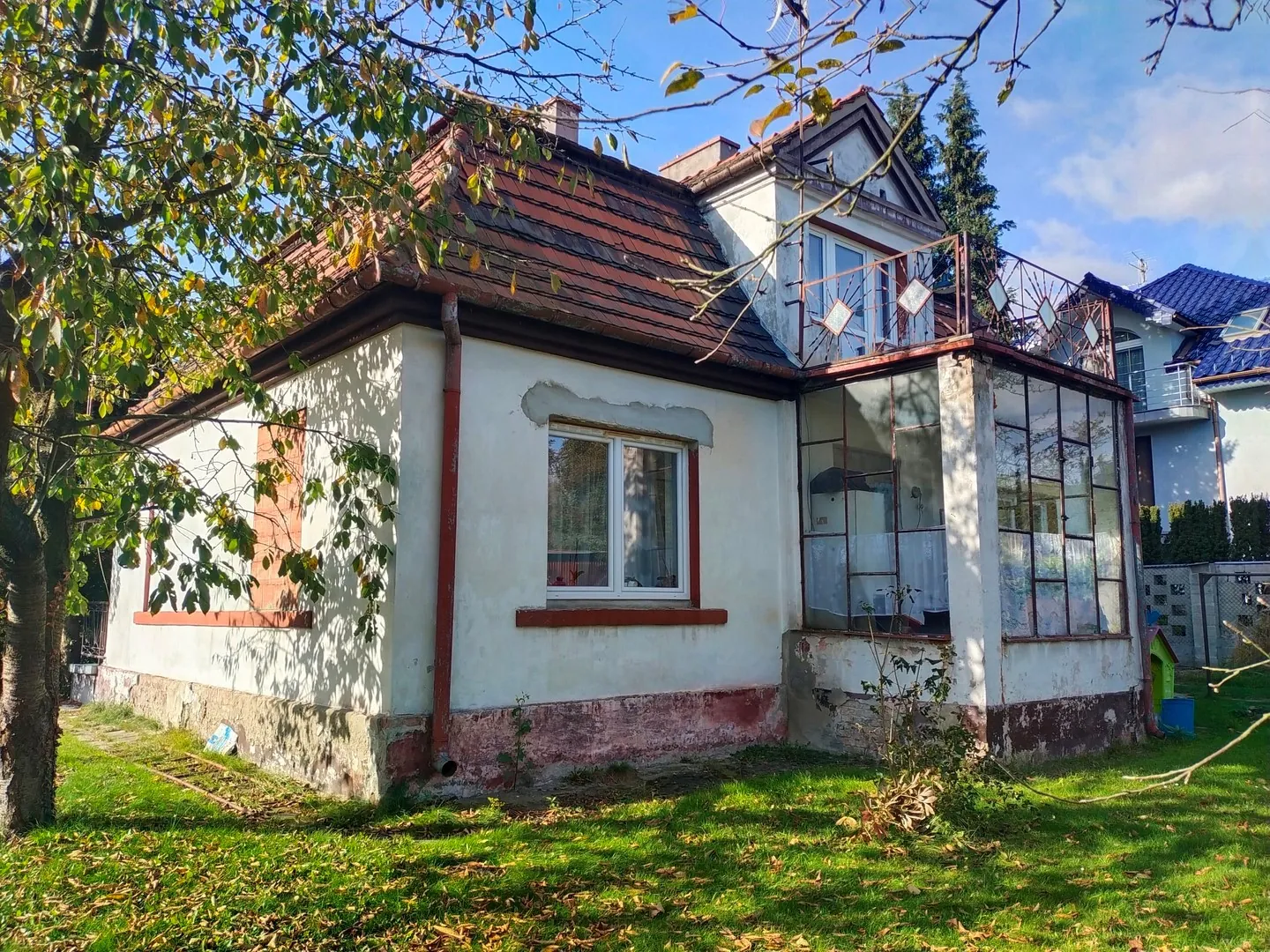 Jednopiętrowy dom z poddaszem i brązowym, dachówkowym dachem. Budynek ma białe ściany z widocznymi śladami zużycia i odpryskami tynku przy fundamencie. Po prawej stronie znajduje się przeszklona weranda oraz balkon na piętrze z metalową balustradą. Przed domem rozciąga się trawnik z opadłymi liśćmi, a po lewej stronie widoczne jest drzewo z zielonymi liśćmi, które częściowo zasłania fasadę.