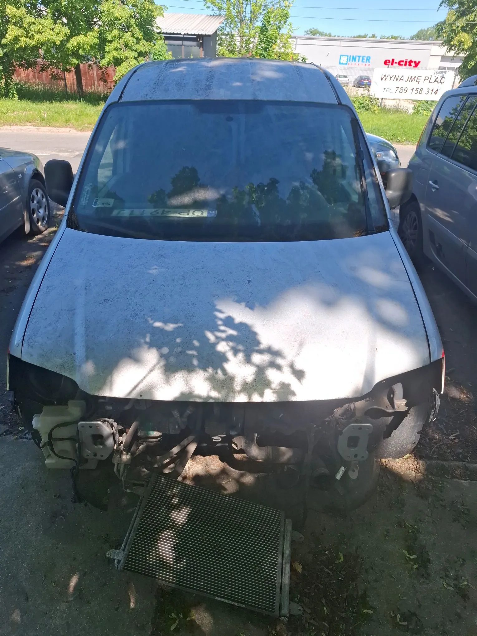 Zdjęcie przodu srebrnego samochodu typu van, który jest uszkodzony. Brakuje przedniego zderzaka oraz grilla, a chłodnica leży na ziemi przed pojazdem. Samochód stoi zaparkowany między innym srebrnym autem a szarym samochodem. W tle widoczny jest biały budynek z napisami: 'INTER', 'el-city' oraz 'WYNAJMĘ PLAC TEL 789 158 314'. Na masce samochodu widoczne są cienie rzucane przez liście drzew.