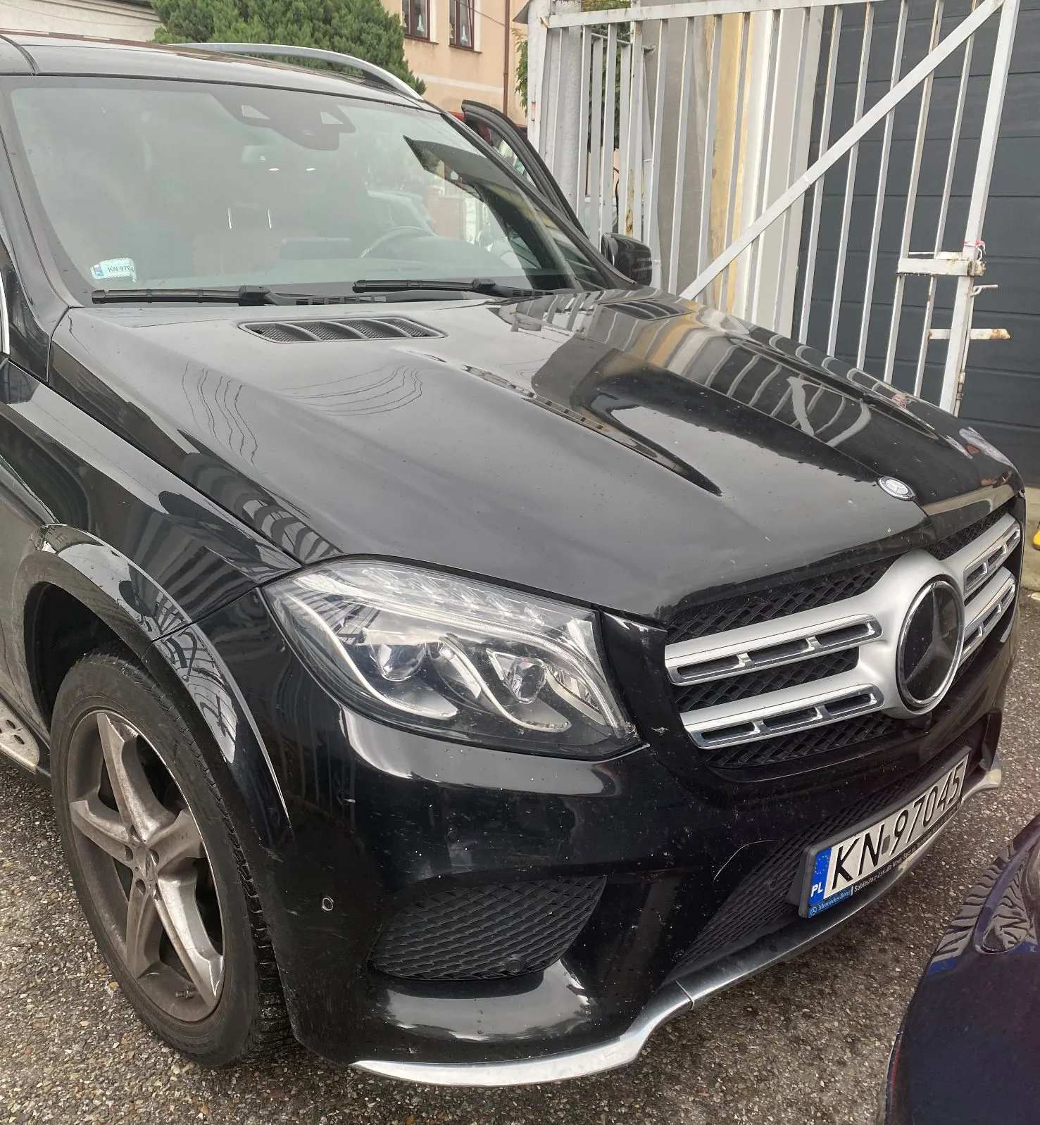 Czarny samochód marki Mercedes-Benz zaparkowany na żwirowym podłożu przed metalową bramą. Widoczny jest przód pojazdu z charakterystycznym logo na grillu, reflektorami oraz tablicą rejestracyjną o numerze KN 9704S.