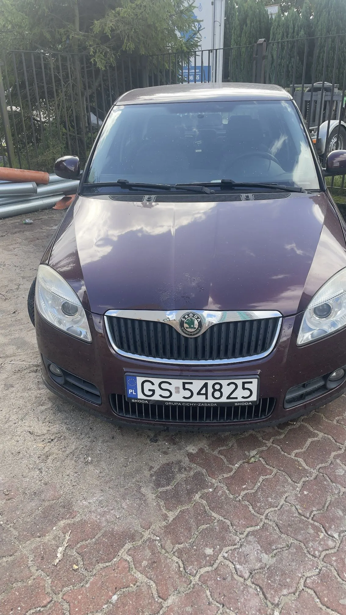 Widok z przodu na bordowy samochód marki Skoda zaparkowany na kostce brukowej. Pojazd posiada polską tablicę rejestracyjną o numerze GS 54825. Na ramce tablicy widnieje napis 'SKODA GRUPA CICHY-ZASADA SKODA'. W tle widać metalowy płot oraz zielone drzewa i krzewy.