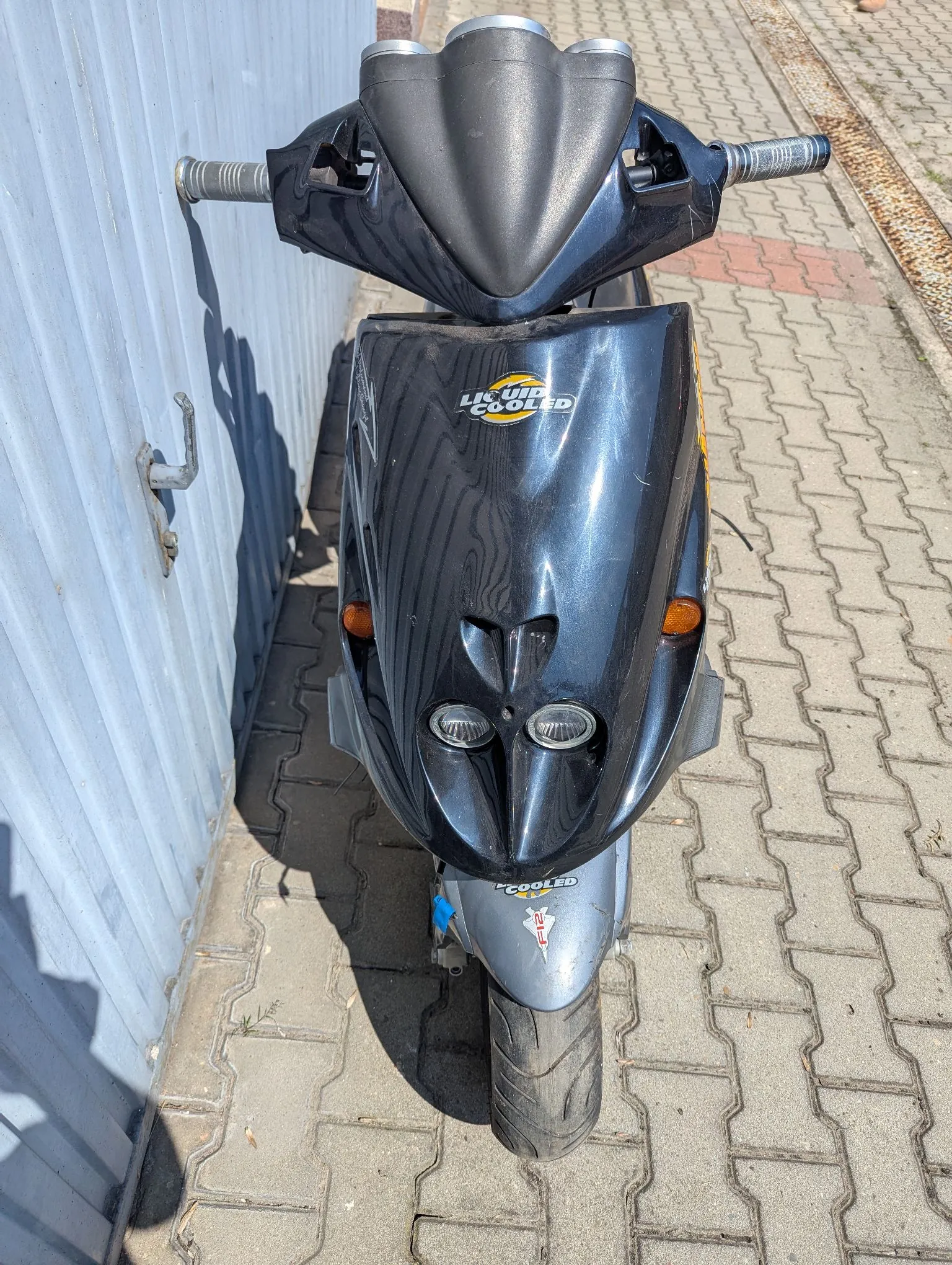 Widok z przodu na czarny skuter zaparkowany na kostce brukowej obok jasnoniebieskiej, falistej ściany. Na przedniej owiewce znajduje się napis „LIQUID COOLED”. Pojazd ma dwa małe reflektory u dołu oraz kierunkowskazy po bokach.
