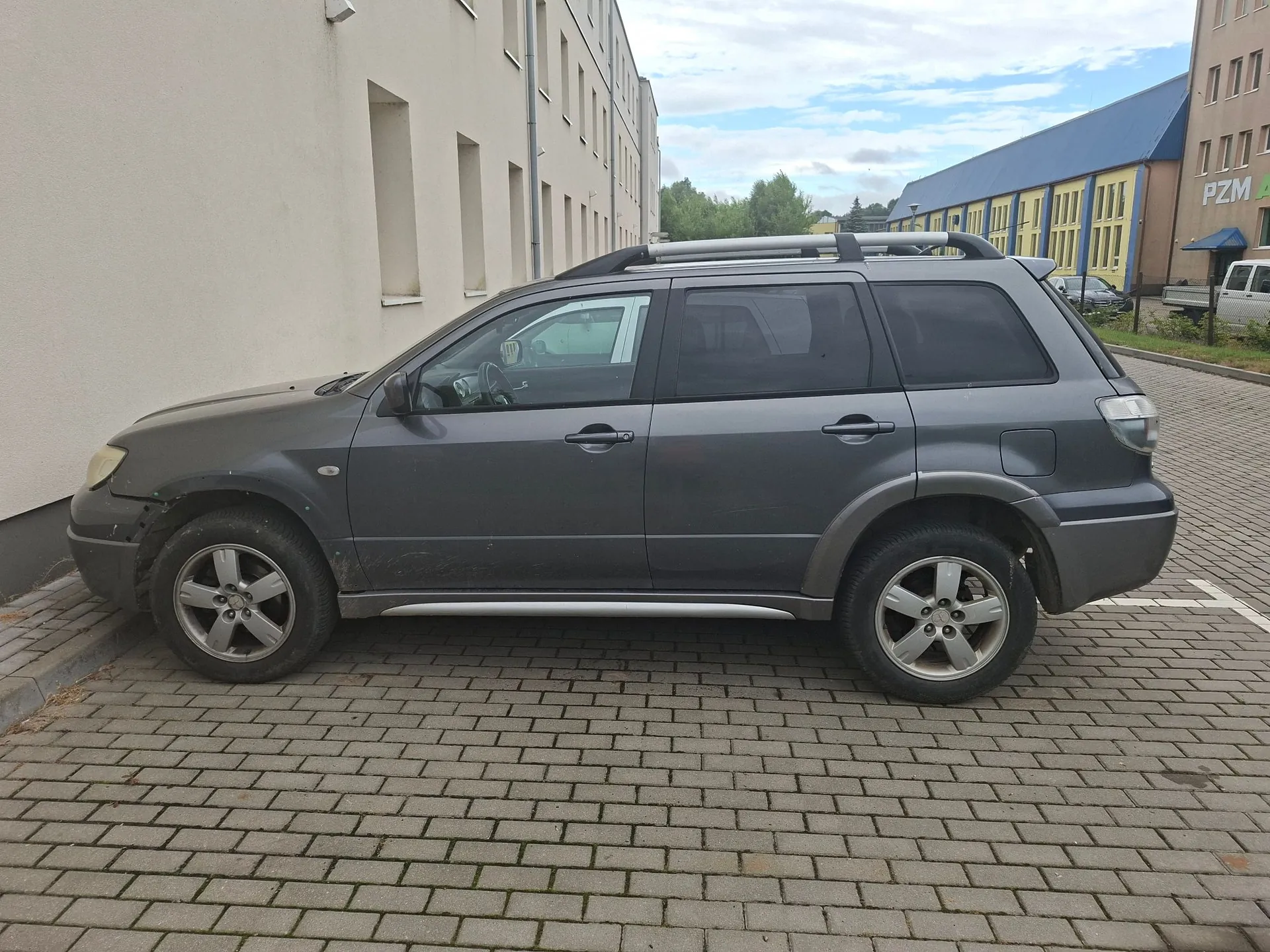 Szary samochód typu SUV zaparkowany bokiem na kostce brukowej przed jasnym budynkiem. W tle widoczny jest niebieski budynek z napisem PZM.