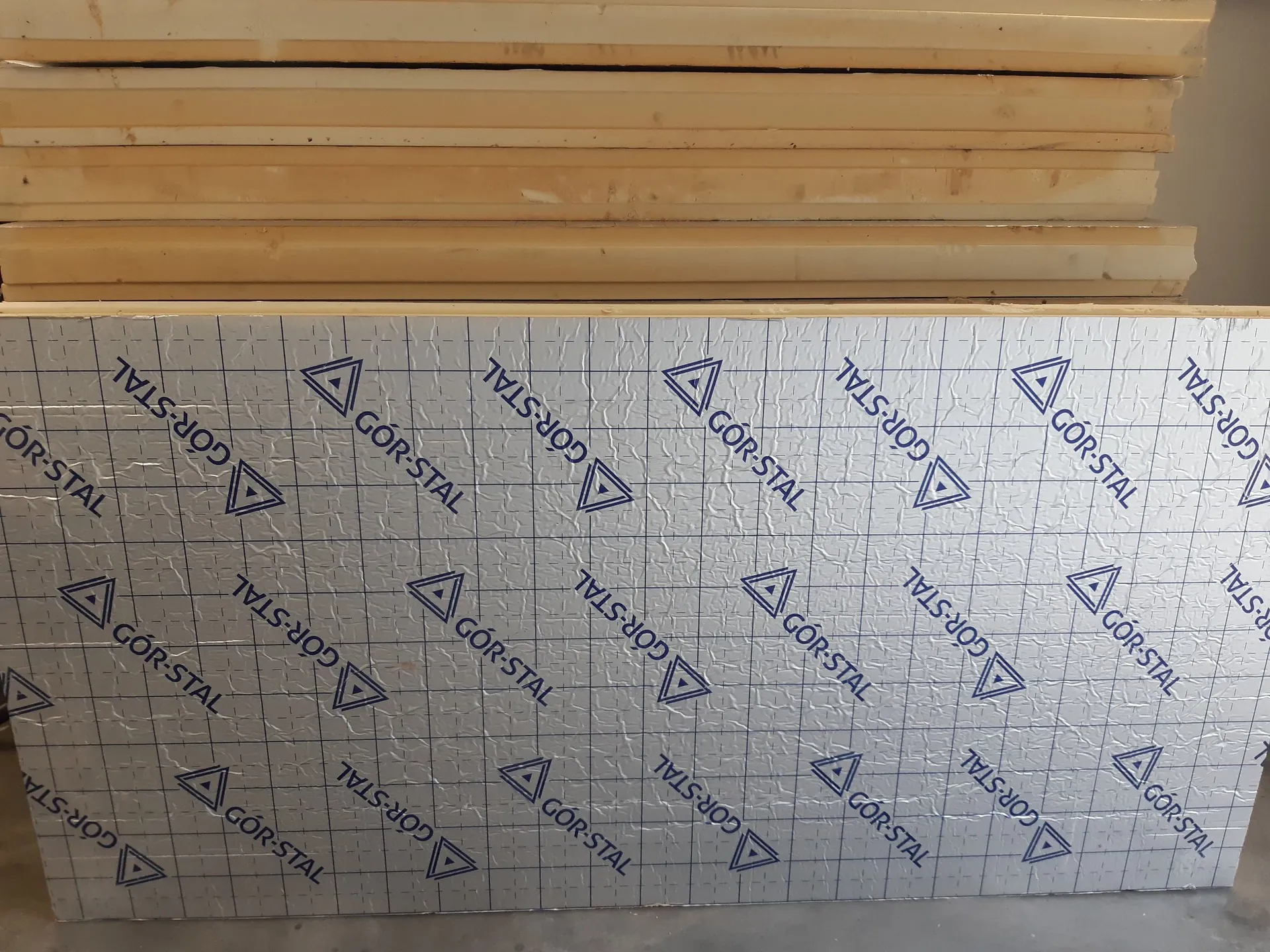 Zdjęcie przedstawiające stos materiałów budowlanych. Na pierwszym planie znajduje się płyta izolacyjna pokryta srebrną folią aluminiową z nadrukowanym powtarzającym się niebieskim logo i napisem „GÖR-STAL”. Powyżej widoczne są ułożone poziomo płyty z jasnobrązowego materiału drewnopodobnego lub gipsowego.