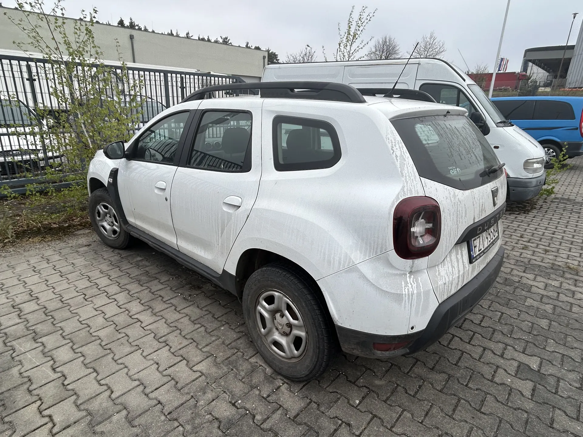 Biały samochód typu SUV (Dacia Duster) z widocznym napisem "Duster" na tylnej klapie, zabrudzony, z tablicą rejestracyjną FZ 9558. Samochód zaparkowany na brukowanej powierzchni, w tle biały i niebieski furgon, metalowe ogrodzenie, budynek oraz drzewa. Niebo zachmurzone.