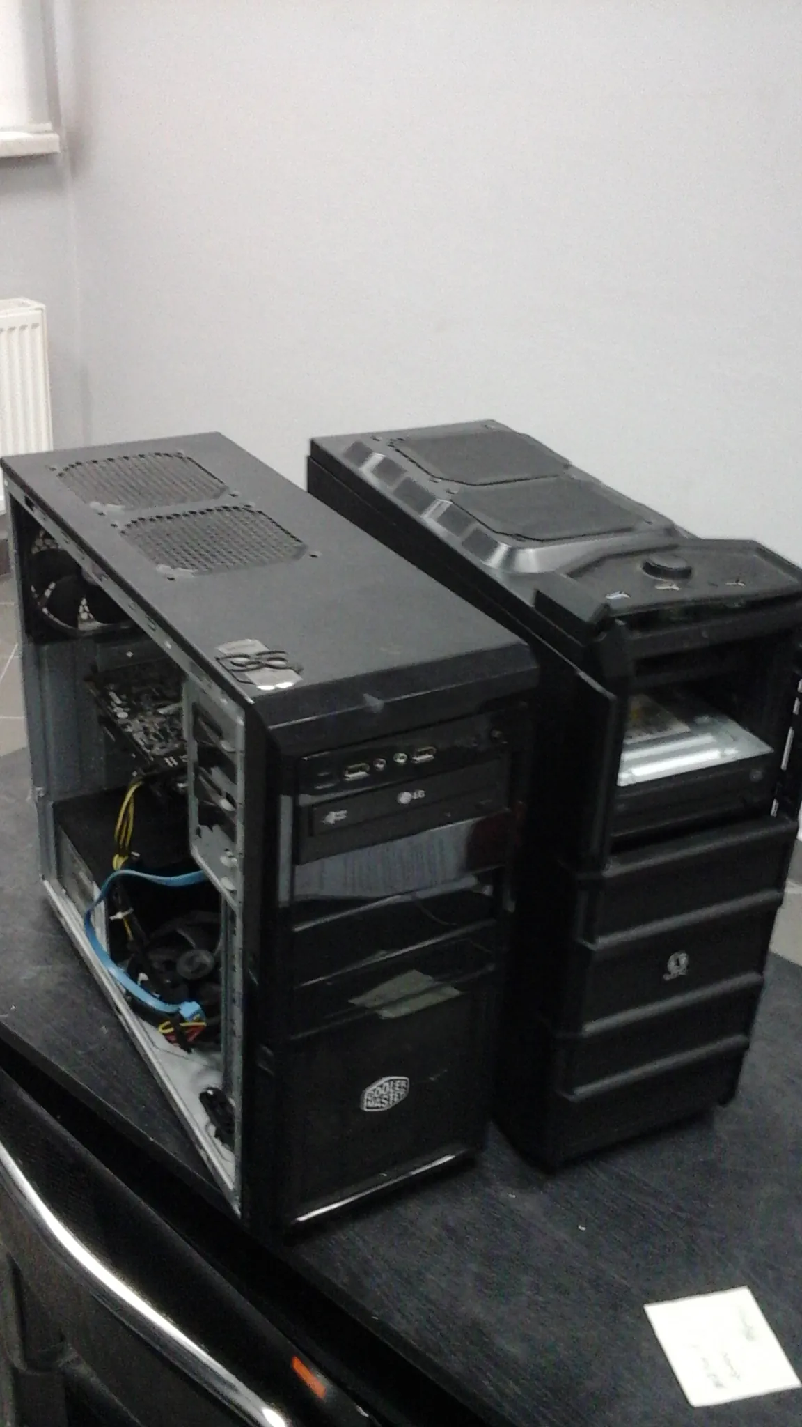 Dwa czarne obudowy komputerowe ułożone na sobie. Dolna obudowa ma zdjętą boczną pokrywę, ujawniając wewnętrzne elementy takie jak przewody (niebieski, żółty), wentylatory i płyta główna. Górna obudowa jest zamknięta, z widocznym logo. Obok nich na ciemnej powierzchni znajduje się biała kartka z nieczytelnym tekstem. Tło stanowi biała ściana i fragment okna.