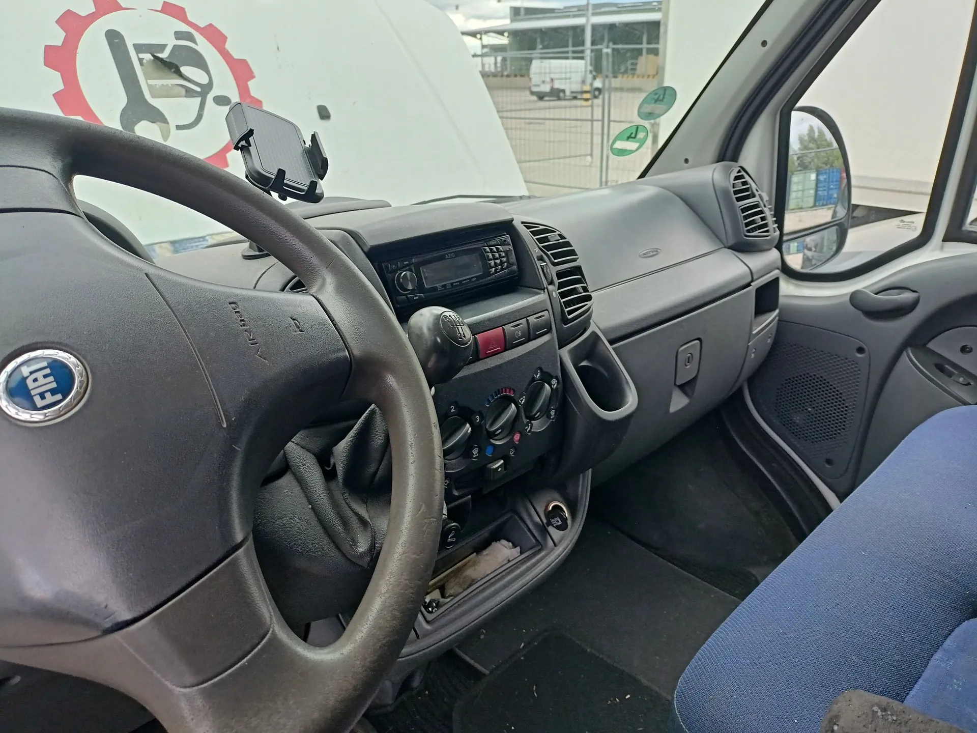 Wnętrze kabiny samochodu marki Fiat, widok z perspektywy kierowcy. W kadrze znajduje się ciemnoszara kierownica z logo FIAT, deska rozdzielcza z radiem, pokrętłami klimatyzacji oraz nawiewami powietrza. Na desce rozdzielczej zamontowany jest uchwyt na telefon. Po prawej stronie widoczny jest fragment niebieskiego fotela pasażera oraz lusterko boczne. Przez przednią szybę widać fragment białego nadwozia z czerwono-białym logo przedstawiającym klucz i tłok w zębatce oraz biały samochód dostawczy w tle.