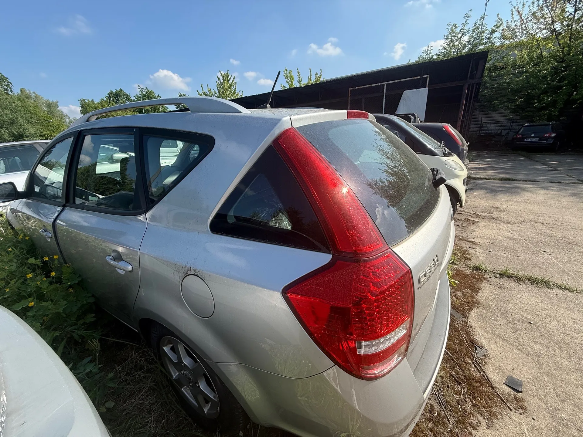Srebrny samochód typu kombi z widocznym napisem "Ceed" na tylnej szybie, zaparkowany na trawie obok betonowego placu. W tle inne pojazdy, budynek z metalowym dachem, drzewa i niebo z kilkoma chmurami. Czerwone światła tylne i srebrne felgi są widoczne. Po lewej stronie rosną rośliny z żółtymi kwiatami.