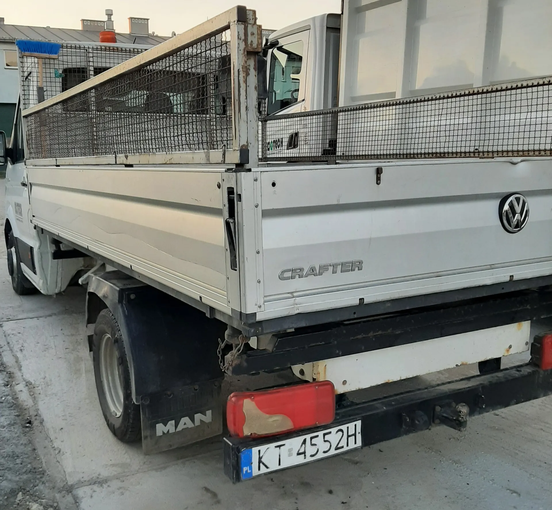 Biały samochód dostawczy Volkswagen Crafter z widocznym tyłem. Na boku napis "CRAFTER" oraz logo VW. Widoczna tablica rejestracyjna PL KT 4552H. Na osłonie koła napis "MAN". Kuzow z metalową siatką, w tle budynki z kominami i niebieska miotła. Samochód stoi na betonowej powierzchni.