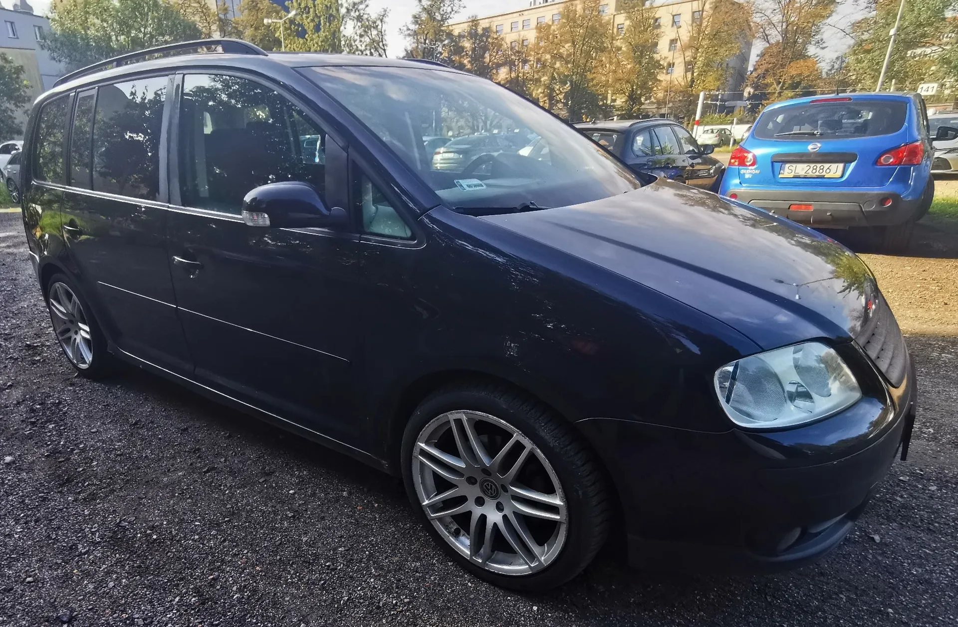 Ciemnoniebieski samochód typu minivan zaparkowany na żwirowym placu. Pojazd jest widoczny z przodu i z boku, posiada srebrne felgi aluminiowe. W tle widoczne są inne zaparkowane samochody, w tym niebieski hatchback z tablicą rejestracyjną SL 2886J, oraz drzewa i budynki mieszkalne.