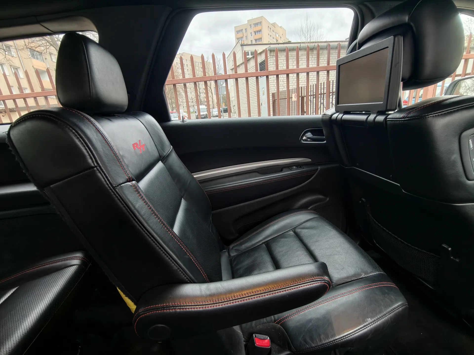 Wnętrze samochodu z czarnymi skórzonymi fotelami z czerwonymi przeszyciami. Na oparciu przedniego fotela widoczny czerwony napis 'R4'. W tle przez okno widać czerwone ogrodzenie, budynki mieszkalne oraz samochody na ulicy. Na oparciu tylnym fotela zamontowany jest ekran. Niebo jest zachmurzone.