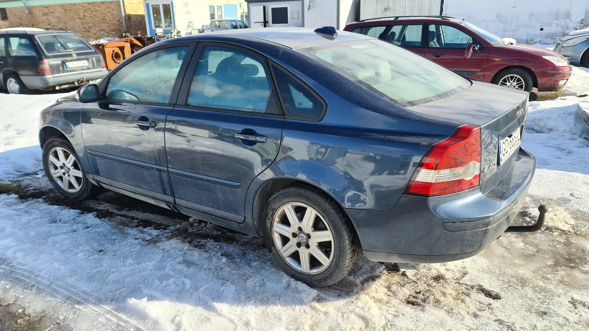 Ciemnoniebieski samochód osobowy typu sedan zaparkowany na śnieżnej nawierzchni. Widoczna tablica rejestracyjna z numerem GD 659L. W tle inne pojazdy: czerwony samochód i szary samochód z tablicą CV 6798. Tło stanowią budynki, w tym budynek z cegły i biały budynek z oknami. Na ziemi śnieg i lodowa nawierzchnia.