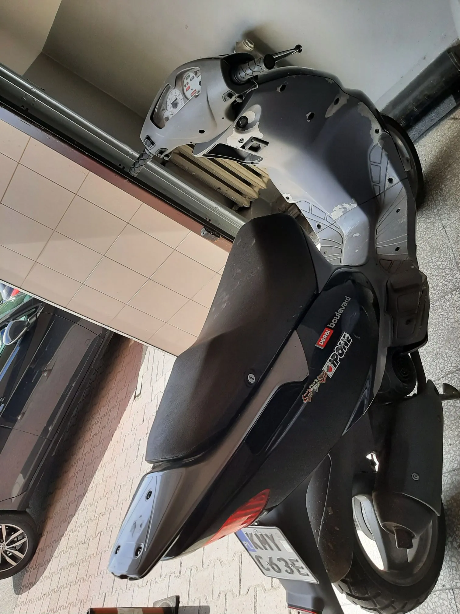 Czarny skuter marki Derbi model Boulevard zaparkowany na chodniku obok budynku. Na boku skutera widnieją naklejki 'IPONE' oraz 'DERBI boulevard'. Pojazd posiada białą tablicę rejestracyjną o numerze 'KWI G63E'. W tle widoczny jest fragment ciemnego samochodu oraz jasna ściana z kafelków.