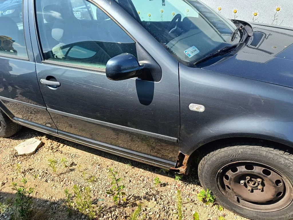 Zbliżenie na prawy bok ciemnoszarego samochodu osobowego. Widoczna jest przednia część drzwi kierowcy, lusterko boczne oraz przednie koło na stalowej feldze. Na dolnej krawędzi progu samochodu, nad przednim kołem, widoczne są ślady korozji i rdzy. Auto stoi na żwirowym podłożu z pojedynczymi kępkami zielonej trawy.