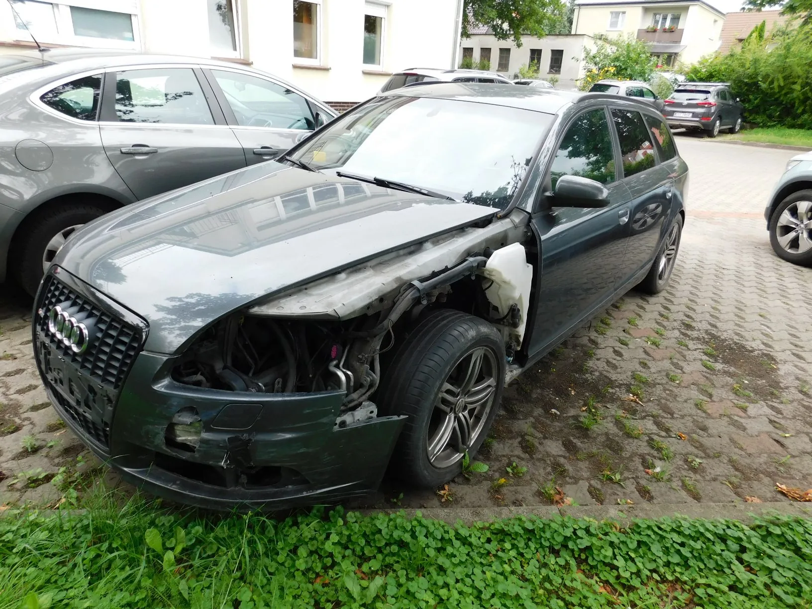 Szary samochód marki Audi w nadwoziu kombi z poważnymi uszkodzeniami przodu po stronie kierowcy. Brakuje reflektora, błotnik jest wgnieciony i częściowo oderwany, a zderzak jest pęknięty i poluzowany. Samochód zaparkowany jest na kostce brukowej obok innego szarego auta. W tle widoczne są białe budynki mieszkalne i zieleń.
