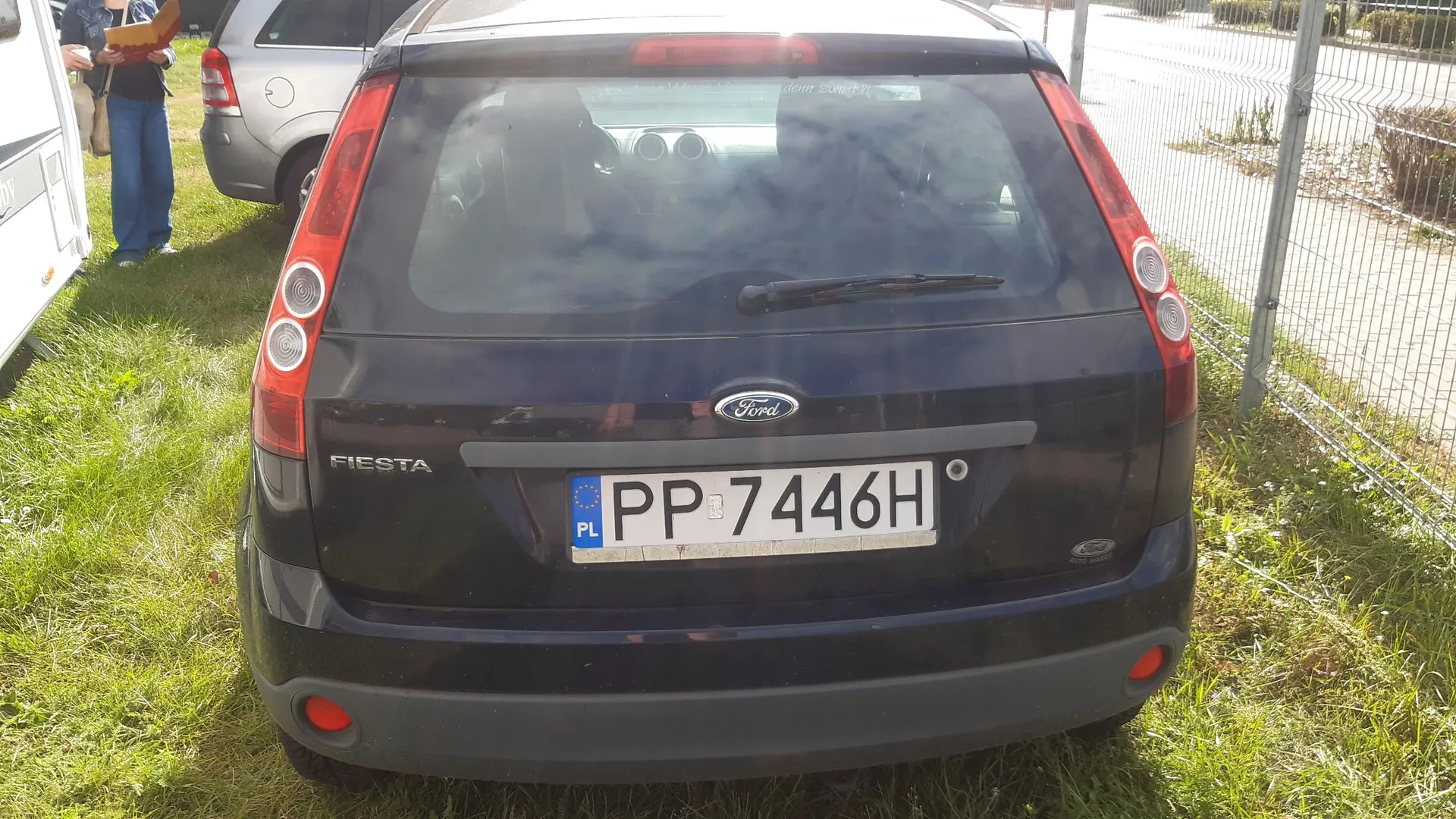 Tylny widok ciemnoniebieskiego samochodu marki Ford Fiesta. Na klapie bagażnika znajduje się logo Ford oraz napis FIESTA po lewej stronie. Samochód ma polską tablicę rejestracyjną o numerze PP 7446H. Pojazd zaparkowany jest na trawie obok ogrodzenia z siatki.
