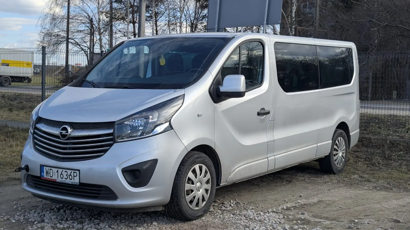 Srebrny samochód dostawczy marki Opel zaparkowany na żwirowym podłożu. Widoczna tablica rejestracyjna z numerem WD 1636P. Pojazd posiada przyciemniane szyby boczne i przednie reflektory. W tle metalowe ogrodzenie, drzewa bez liści, droga oraz żółty ciężarówka. Niebo częściowo zachmurzone.