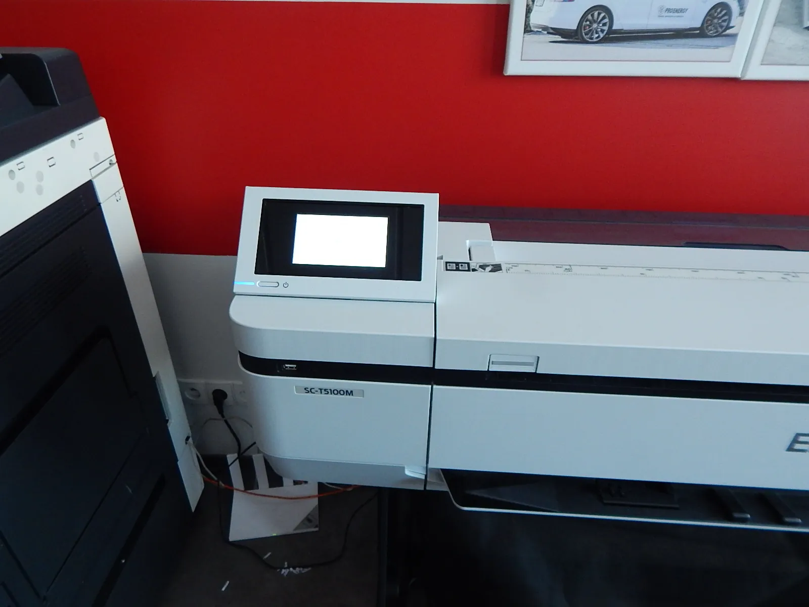 Biała drukarka wielkoformatowa z panelem sterowania z ekranem LCD. Na obudowie urządzenia widnieje napis 'SC-T5100M'. Drukarka stoi na ciemnym blacie na tle czerwonej i białej ściany. Po lewej stronie widoczny jest fragment innego, czarnego urządzenia biurowego.