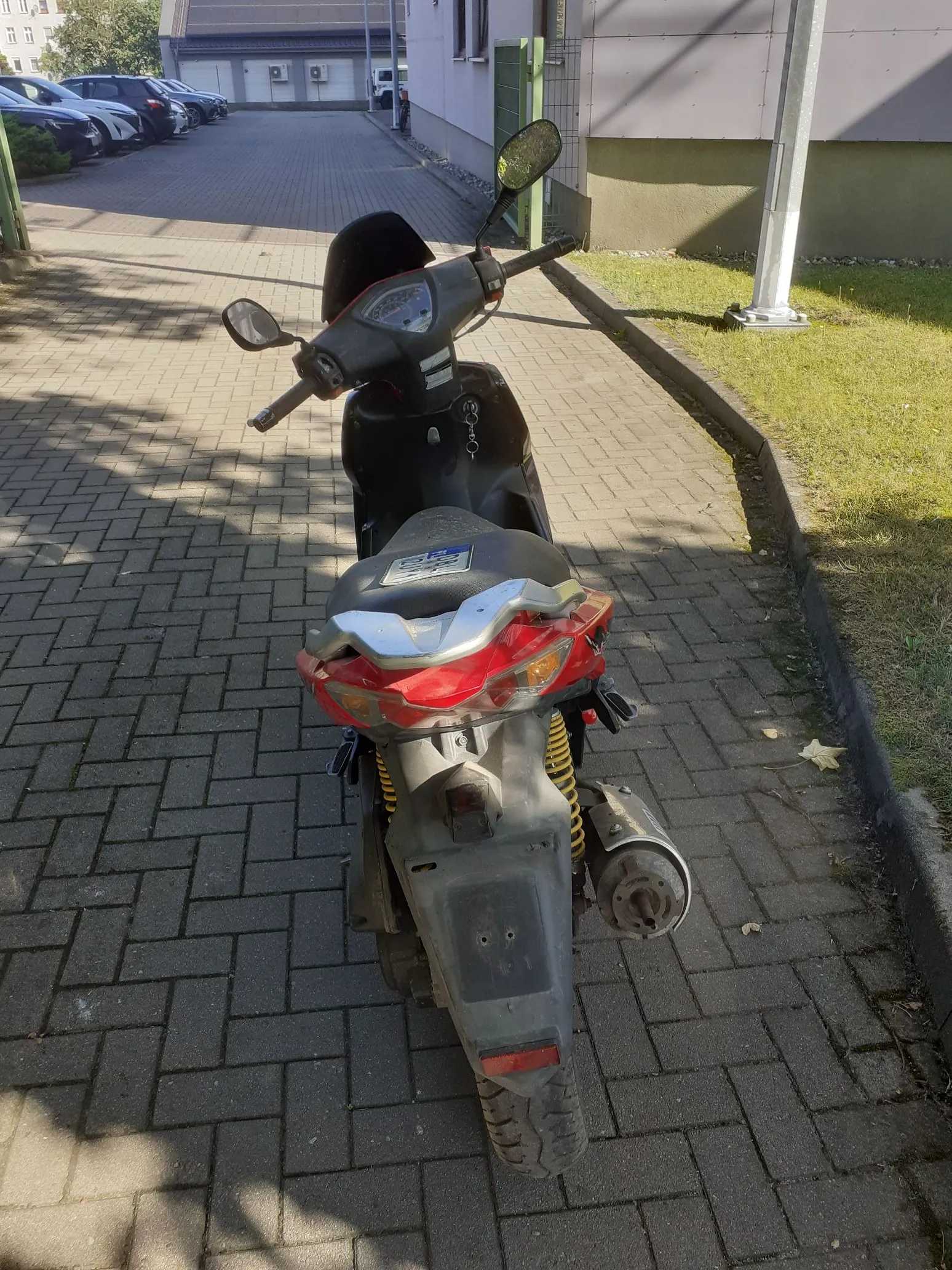 Widok z tyłu na ciemny skuter zaparkowany na kostce brukowej. Pojazd ma czerwone tylne światło, srebrny wydech oraz widoczną tablicę rejestracyjną. W tle widać fragment trawnika, chodnik oraz budynek.