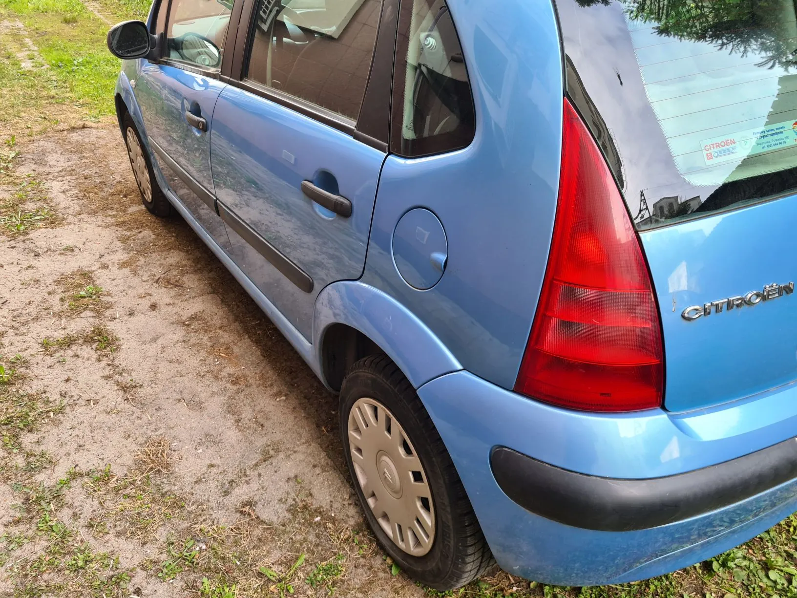 Niebieski samochód marki Citroën zaparkowany na brudnej, częściowo zarośniętej drodze. Widoczne są czerwone światło tylne, pokrywa zbiornika paliwa oraz napis 'CITROËN' na tylnej części pojazdu. W tle trawa i fragment drogi.
