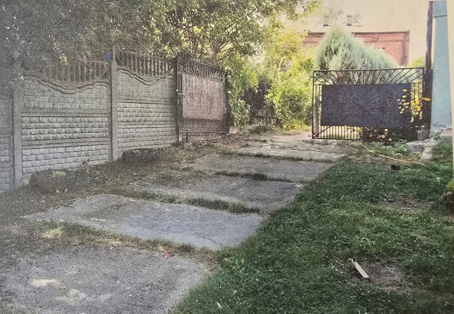 Zdjęcie przedstawiające podwórze z betonową nawierzchnią ułożoną z dużych płyt. Po lewej stronie znajduje się wysoki, szary betonowy płot o dekoracyjnym wzorze, za którym rosną drzewa. W głębi widoczna jest ciemna, metalowa brama oraz zielone krzewy i drzewa. Po prawej stronie znajduje się pas trawnika.